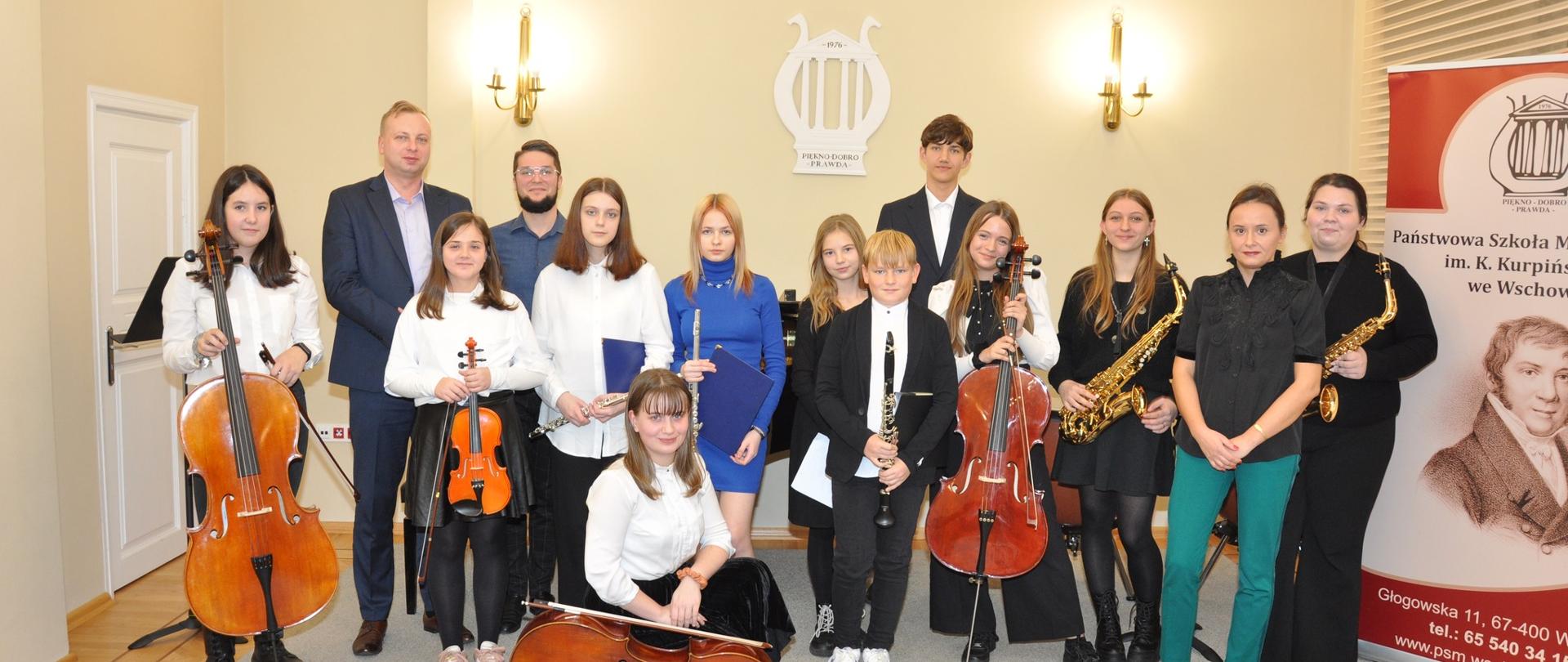 Dziesięcioro uczniów z instrumentami oraz nauczyciele. W tle na ścianie logo szkoły. Z prawej strony baner z nazwą, logo oraz wizerunkiem patrona szkoły.