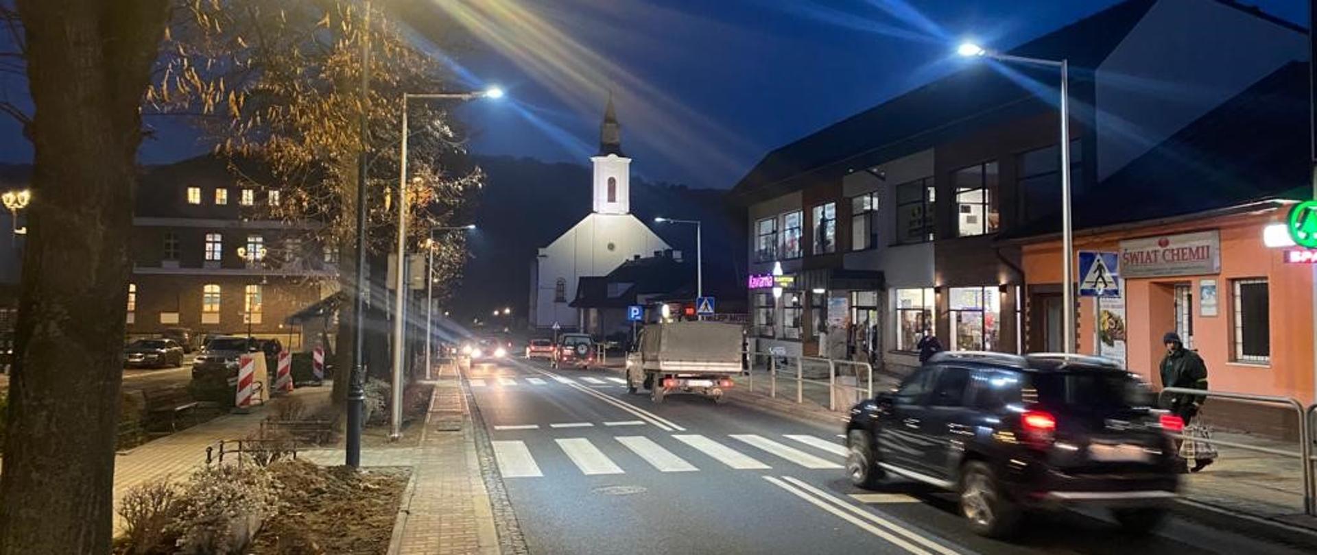 droga w centrum miejscowości, po lewej drzewa, w środku przejście dla pieszych, w tle kościół