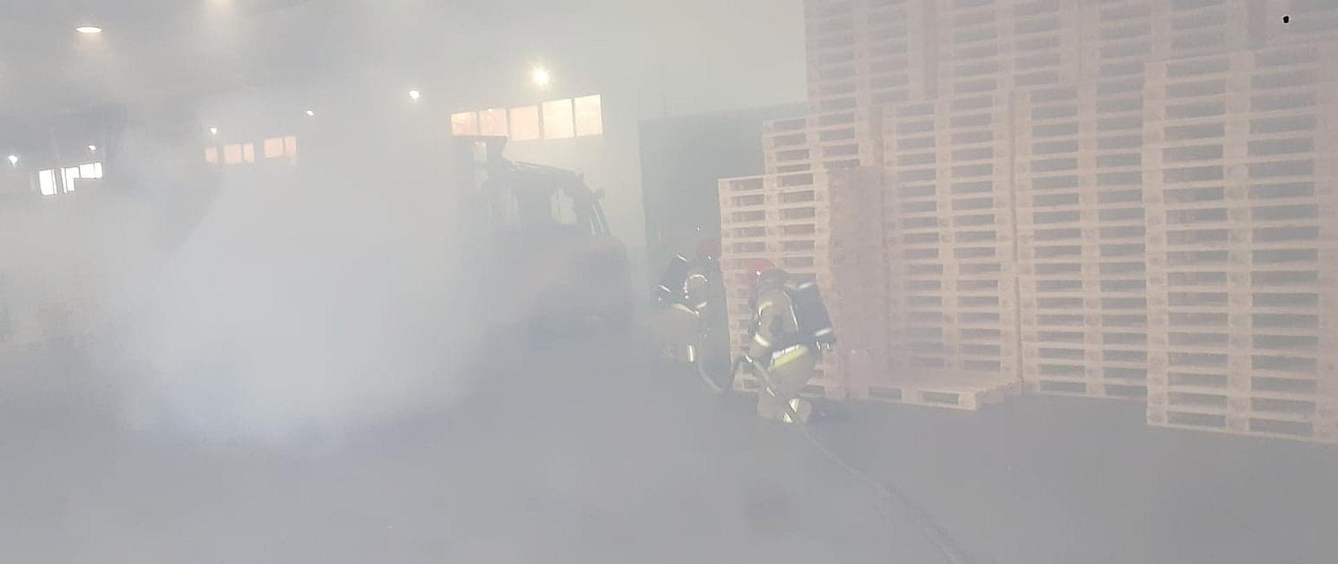 Strażacy ubrani w aparaty OUO idą z linią gaśniczą przez halę na której znajdują się palety. Obiekt jest zadymiony.
