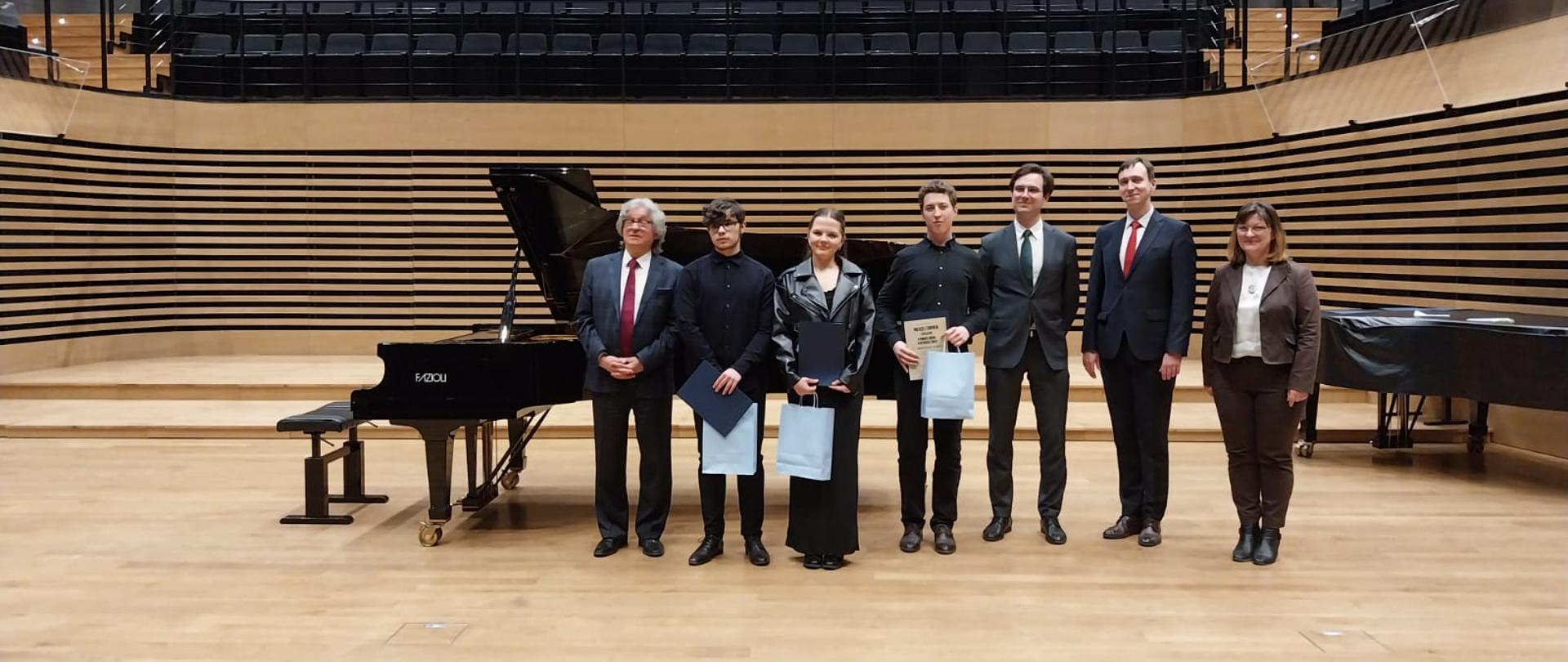 Na scenie stoją w rzędzie: dziewczyna i dwóch chłopców a dyplomami, obok nich trzech mężczyzn i kobieta.