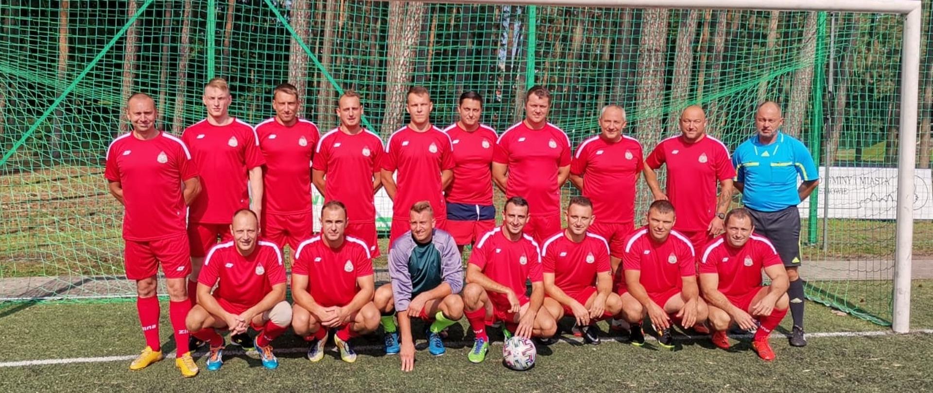 Zdjęcie grupowe przedstawia funkcjonariuszy ubranych w czerwone stroje piłkarskie ustawionych na tle bramki piłkarskiej.