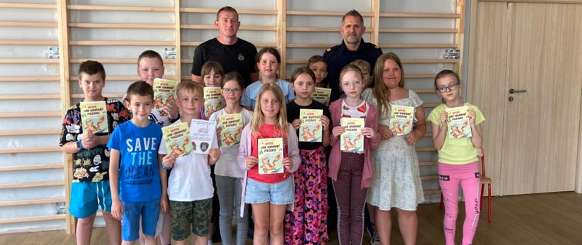Zdjęcie przedstawia dzieci w wieku szkolnym oraz strażnika miejskiego i strażaka. Wszyscy stoją na tle drabinek gimnastycznych w sali sportowej. Dzieci trzymają w ręku broszury informacyjne.