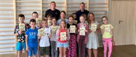 Zdjęcie przedstawia dzieci w wieku szkolnym oraz strażnika miejskiego i strażaka. Wszyscy stoją na tle drabinek gimnastycznych w sali sportowej. Dzieci trzymają w ręku broszury informacyjne.