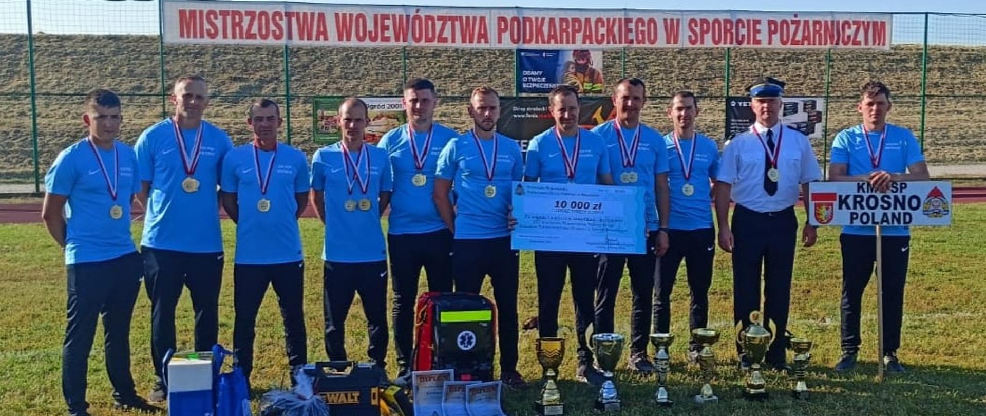 Na zdjęciu widzimy reprezentację Komendy Miejskiej Państwowej Straży Pożarnej w Krośnie wraz z Komendantem Miejskim st. bryg. Zbigniewem Nowakiem. Ustawieni są w jednym szeregu na murawie stadionu. Na szyjach powieszone mają medale. Przed nimi znajdują się zdobyte puchary oraz nagrody rzeczowe m. in. elektronarzędzia oraz torba R1. Jeden ze strażaków trzyma w rękach symboliczny czek na kwotę 10 000 zł jako nagrodę finansową dla drużyny za zwycięstwo w klasyfikacji generalnej.