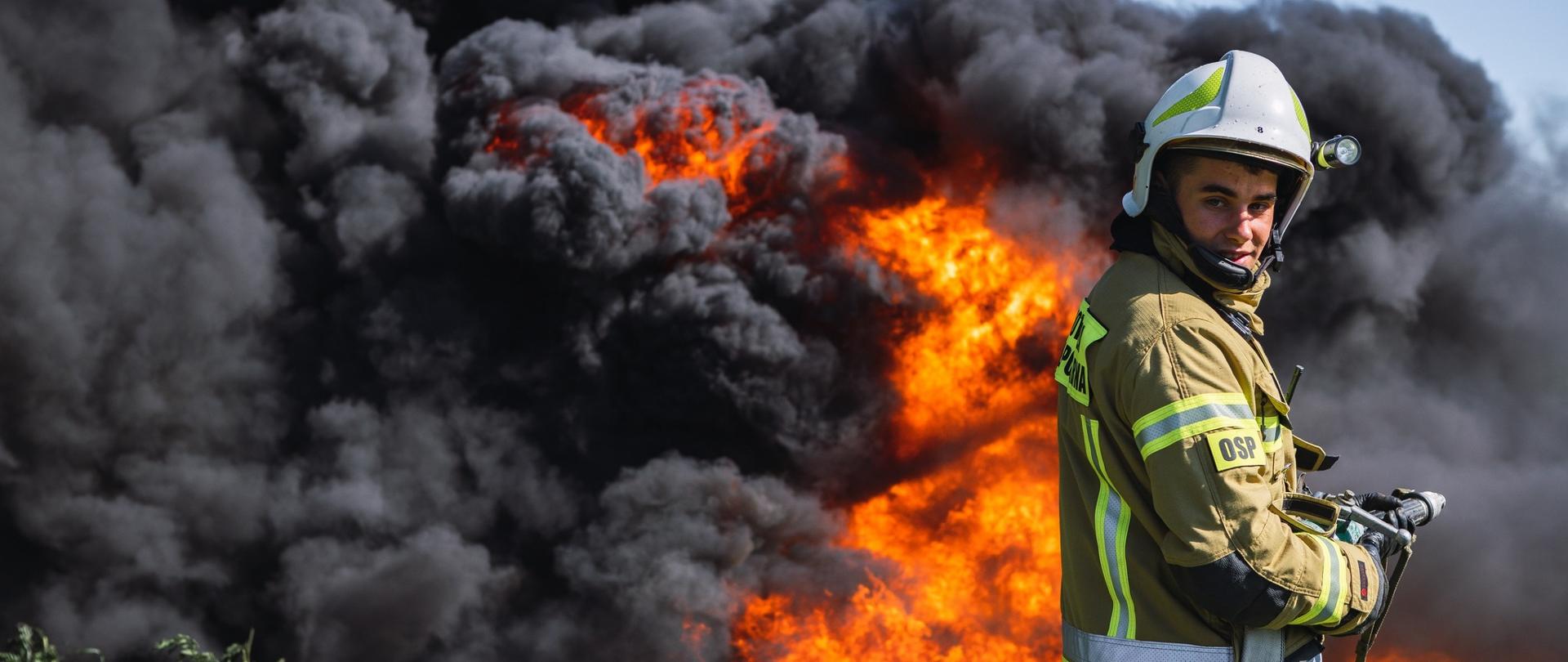 Zdjęcie przedstawia strażaka w umundurowaniu specjalnym. W tle pożar - duża ilość czarnego dymu. 