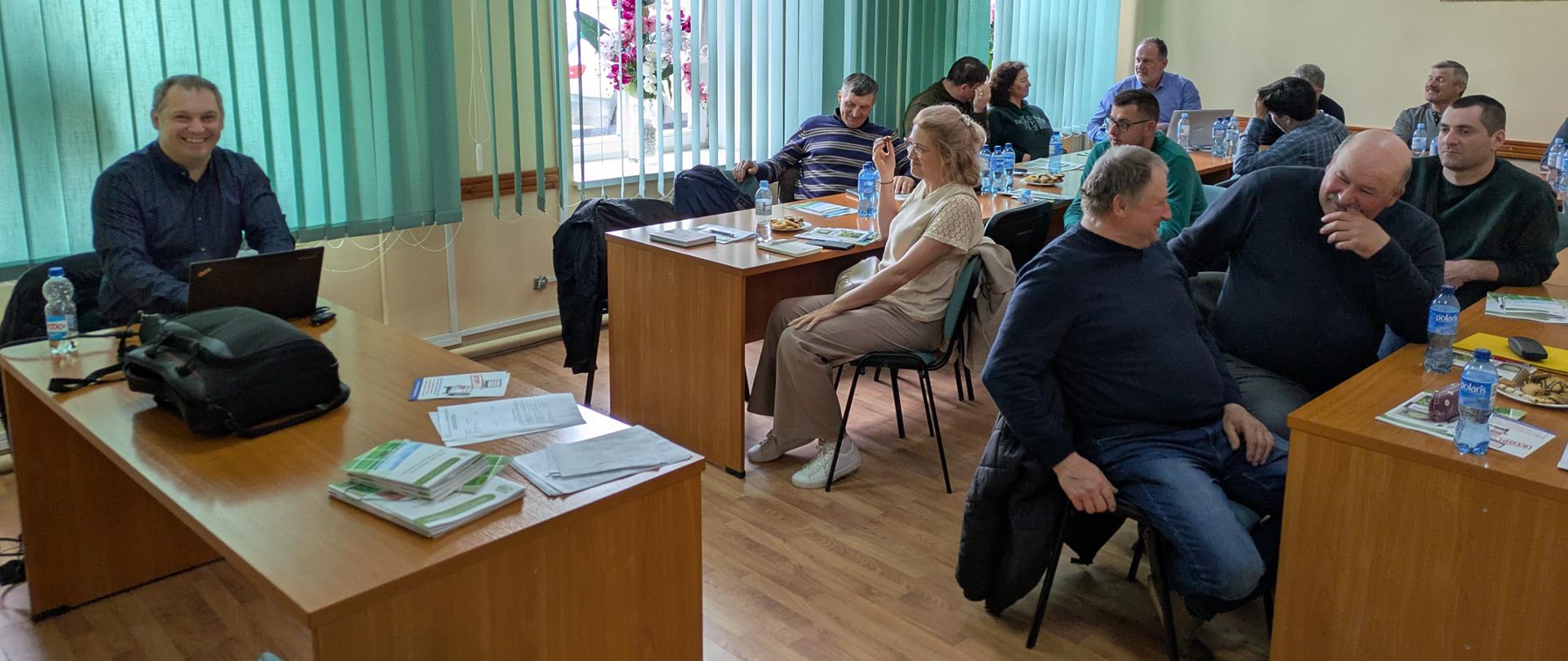 Ludzie siedzą przy drewnianych stołach. Za ludźmi zielona zasłona i żółte ściany