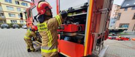 Zdjęcie przedstawia strażaków sprawdzających sprzęt techniczny pożarniczy.
W tle budynek Komendy i pojazd pożarniczy.
