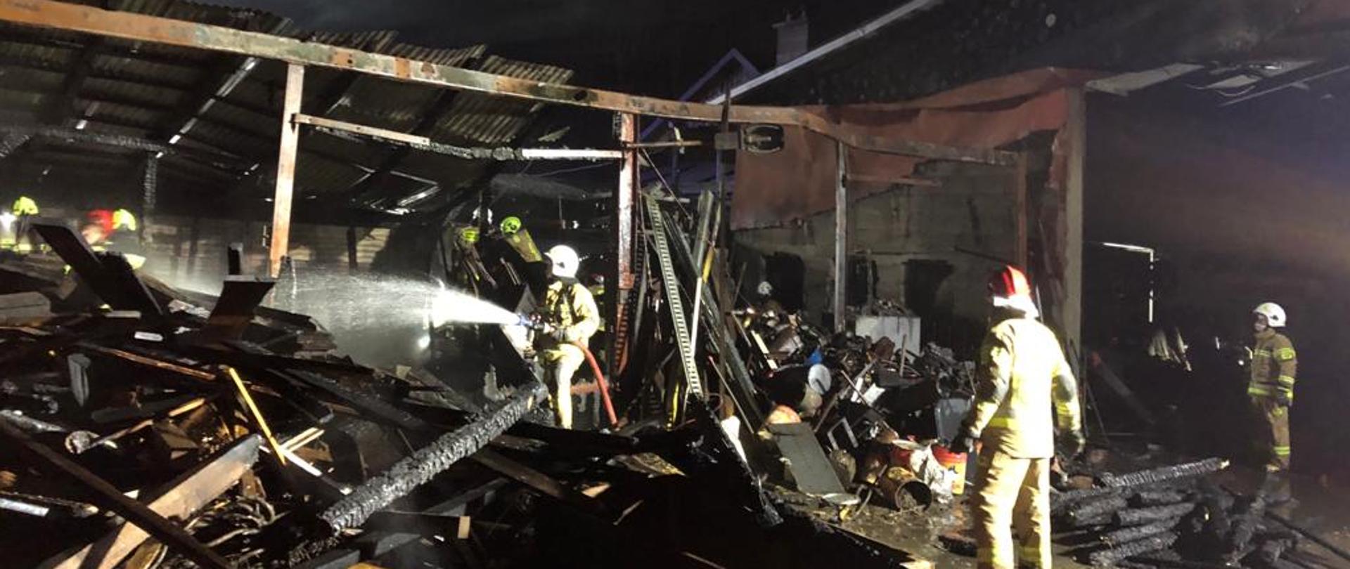 Zdjęcie przedstawia pogorzelisko po pożarze budynku gospodarczego. Wśród zgliszcz przemieszczają się ratownicy demontując spalone elementy konstrukcji.