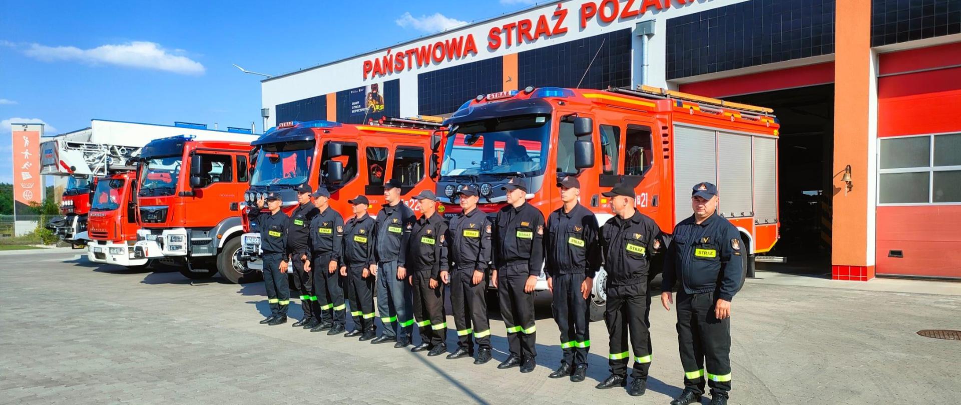 Strażacy stoją w szeregu na placu przed komendą na tle samochodów strażackich.
