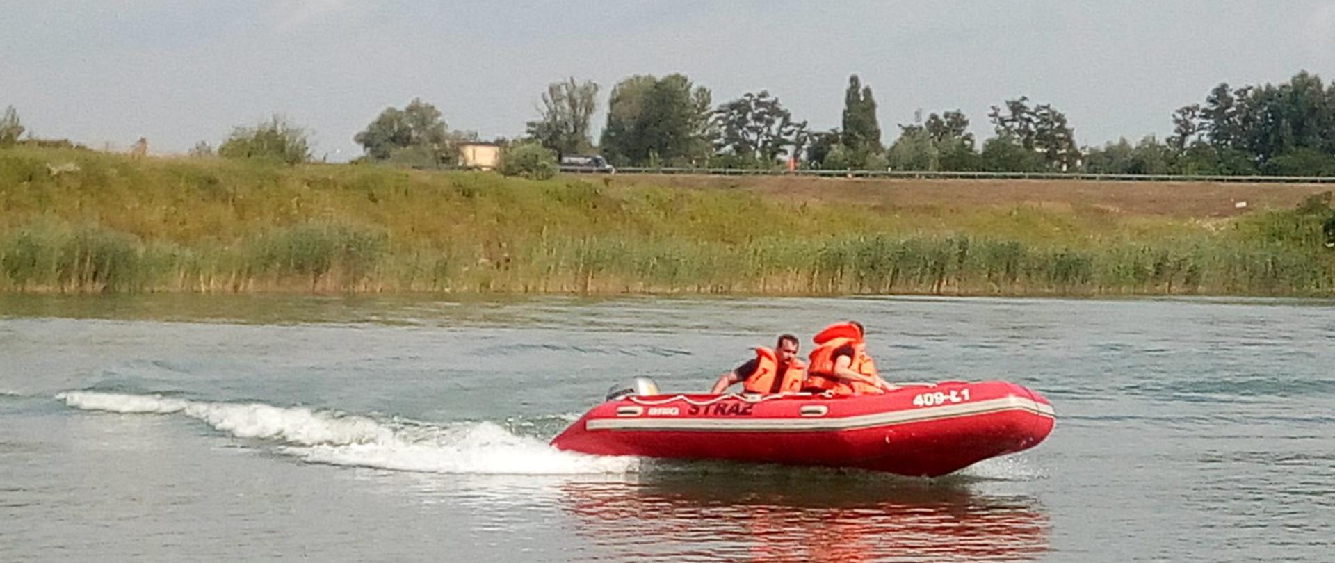 Zdjęcie ukazuje dwóch strażaków płynących pontonem po akwenie wodnym. Dwóch ratowników ubranych jest w pomarańczowe kamizelki asekuracyjne. 