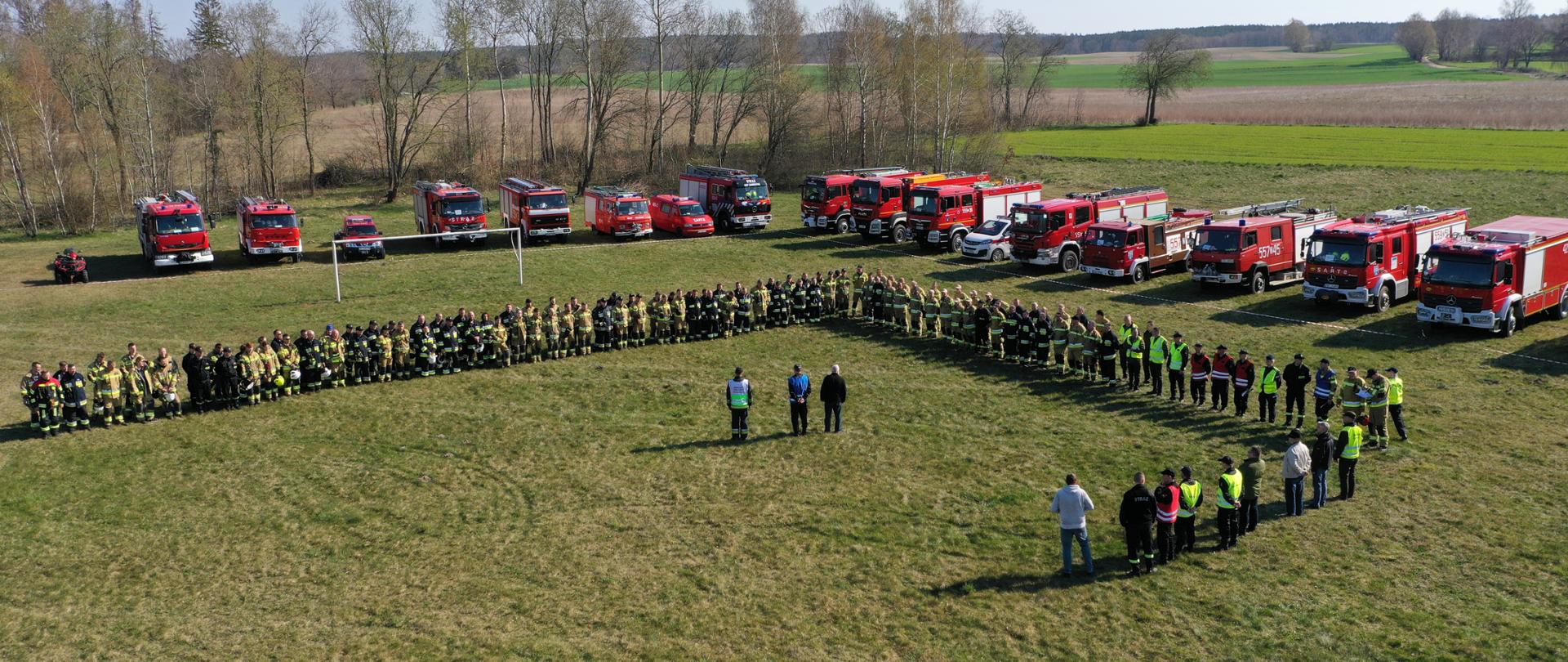 Zdjęcie przedstawia miejsce zbiórki przed Ćwiczeniami LASY 2022. Strażacy ustawieni w dwuszeregu słuchają kierujące działaniem ratowniczym. W tle samochody strażackie oraz lasy i pola.
