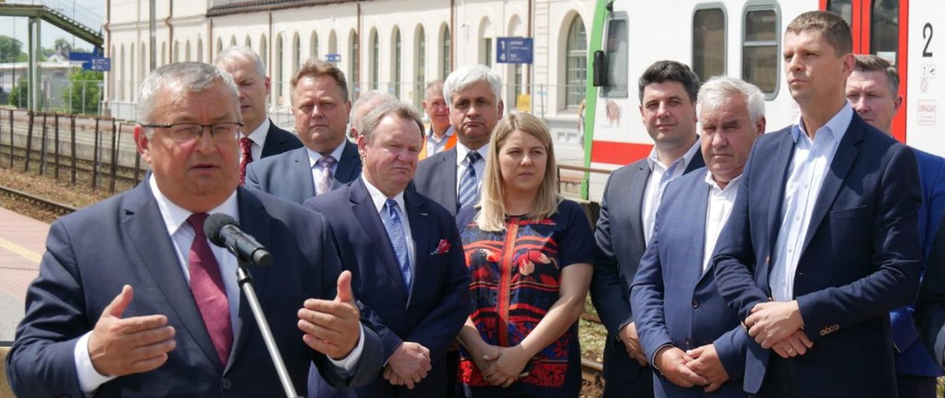 Na zdjęciu widać grupę ludzi. Wszyscy stoją na na peronie kolejowym, w tle widać biały pociąg z czerwonym drzwiami. W tle jest budynek dworca kolejowego. 