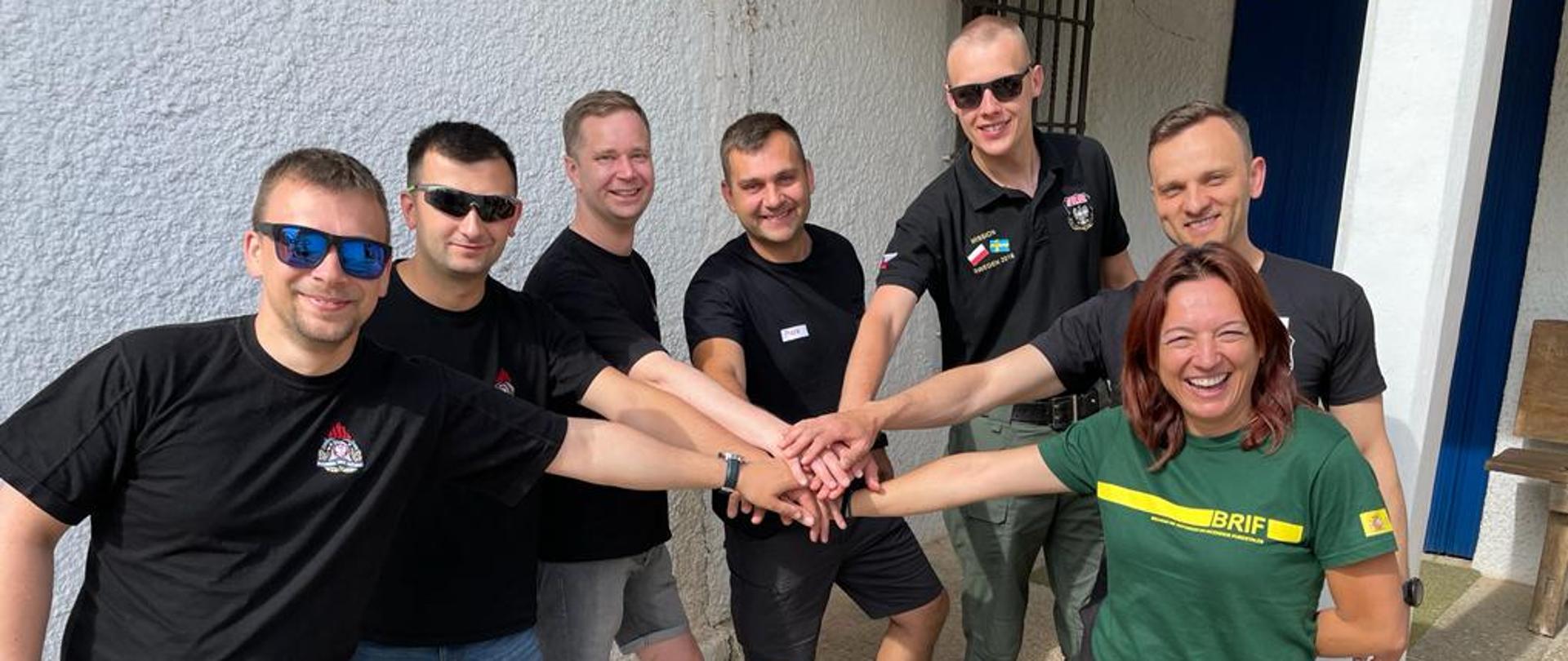 Grupa 6 mężczyzn i kobieta stoją w kole podając sobie nawzajem ręce do środka