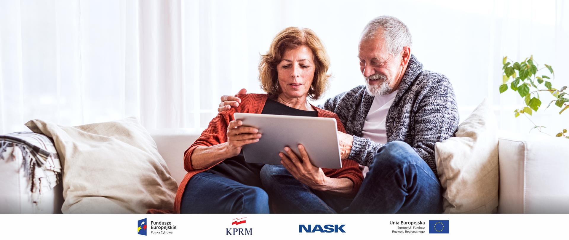 Na zdjęciu widać kobietę i mężczyznę w wieku około 60 lat. Siedzą na jasnej kanapie, są uśmiechnięci i wspólnie korzystają z tabletu. Ubrani są w jeansy i luźne swetry. W tle duże okno przysłonięte jasnymi zasłonami a obok kanapy widać zielone liście kwiatów doniczkowych. 