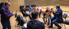 Uczeń i prowadzący warsztaty grający na gitarze oraz czwórka uczniów i nauczyciel przysłuchujący się na scenie sali koncertowej PSM