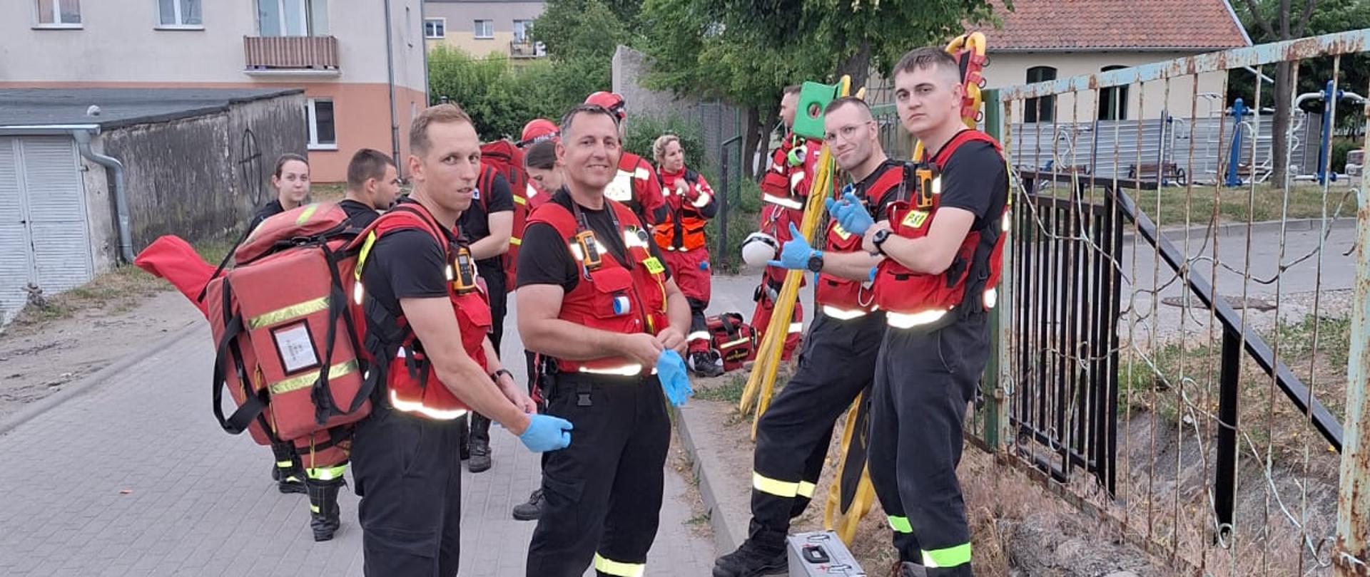 grupa strażaków w czerwonych kamizelkach z torbą medyczną i deską ratowniczą stoją przy płocie