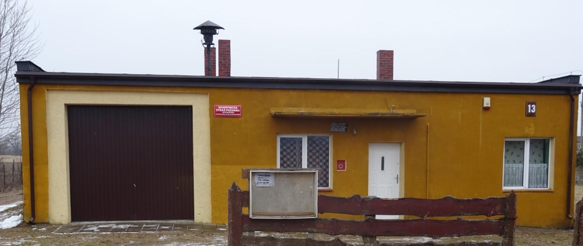 Budynek parterowy koloru żółtego z bramą garażową koloru brązowego. Na środku budynku wejście główne. Podjazd do bramy garażowej z utwardzonej nawierzchni ziemi. 