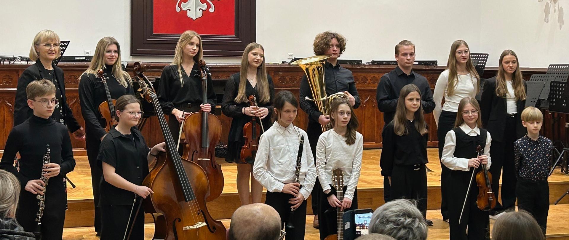Grupa uczniów klasy II PSM I stopnia, cyklu 4-letniego, stoi na scenie Sali Królewskiej z instrumentami, m.in. skrzypcami, wiolonczelami, kontrabasem, klarnetami i tubą; z lewej strony stoi nauczycielka odpowiedzialna za organizację koncertu.
