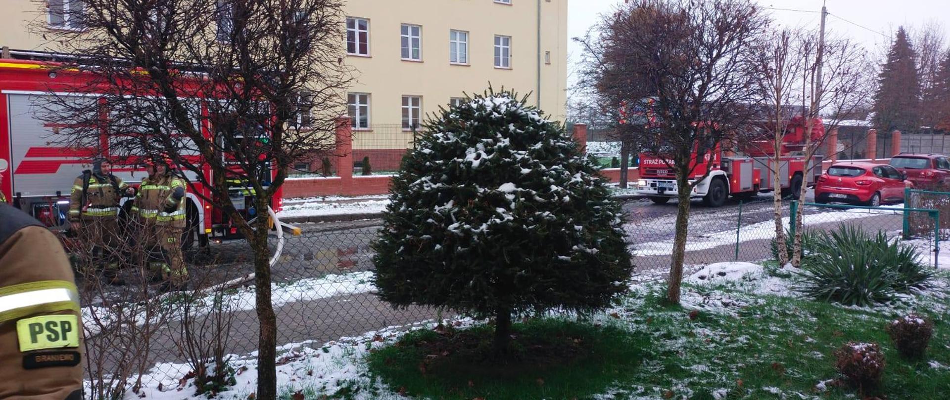 Na zdjęciu z prawej i lewej strony na drodze ustawione samochody strażackie. Na dole zielona trawa pokryta częściowo śniegiem. W oddali duży budynek biurowy.