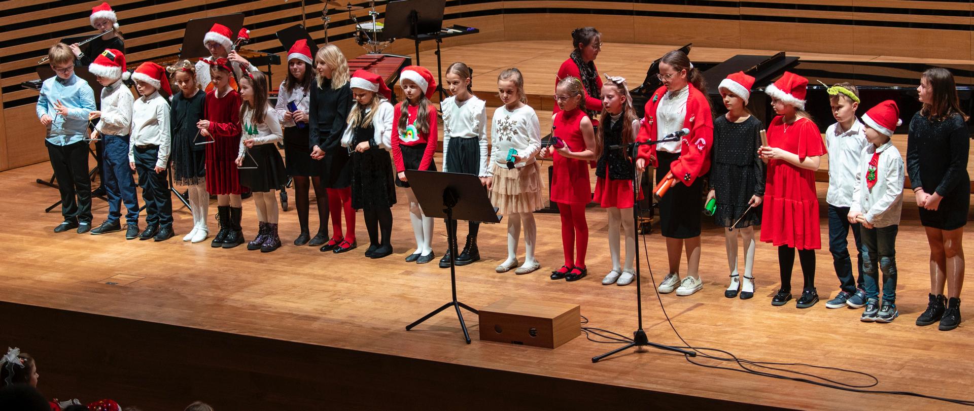 Duża grupa dzieci stoi w rzędzie i śpiewa, za nimi kobieta akompaniuje na fortepianie, chłopiec gra na wiolonczeli, dziewczynka na flecie.