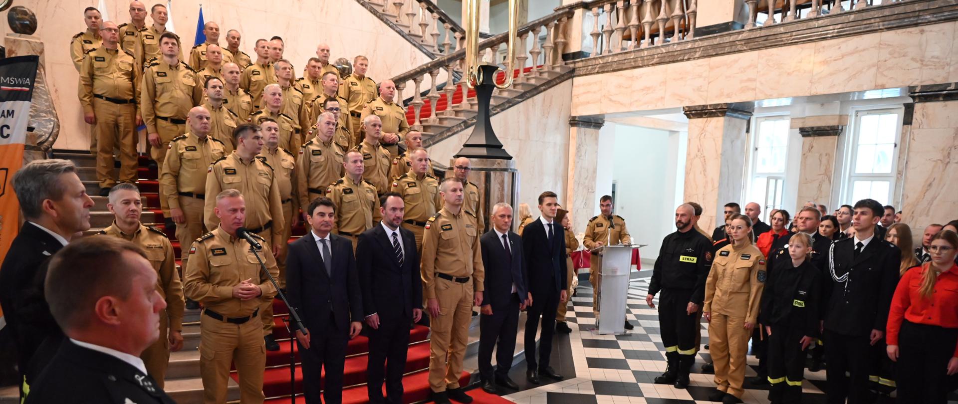 Uroczysty apel odbywający się w eleganckim holu budowli z marmurowymi elementami i czarno-białą posadzką. Na szerokich schodach przykrytych czerwonym dywanem stoi w szyku grupa kilkudziesięciu strażaków Państwowej Straży Pożarnej w piaskowych mundurach służbowych. Przed schodami ustawieni są przedstawiciele władz oraz kadry kierowniczej w garniturach i mundurach, a jeden ze strażaków przemawia do mikrofonu. Po prawej stronie znajdują się pozostali uczestnicy wydarzenia, w tym strażacy oraz druhowie w czarnych mundurach galowych.