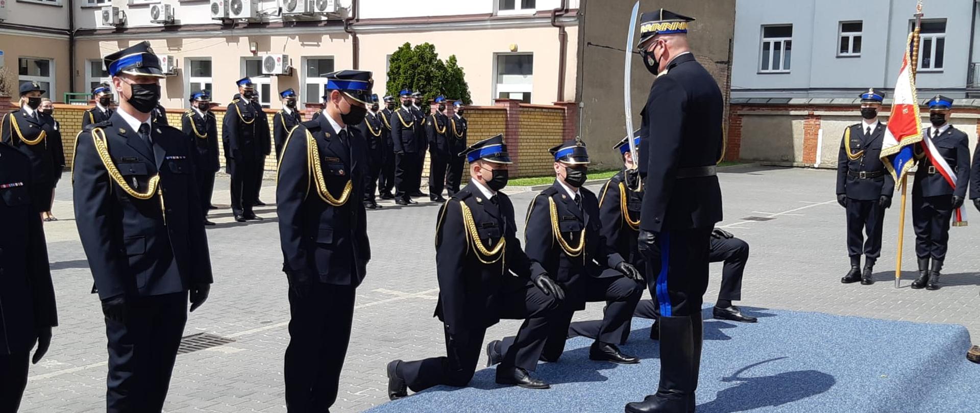 Zastępca Komendanta Głównego PSP dokonuje aktu promocji oficerskiej szablą, którą trzyma w prawej ręce, przed nim funkcjonariusze klęczą na prawym kolanie