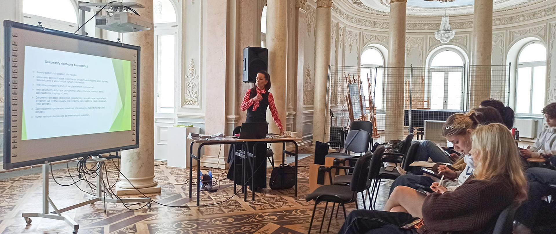 Ujęcie z prawej strony sali. Prowadząca stoi przy ekranie i gestykuluje prawą ręką, tłumacząc treść slajdu zatytułowanego „Dokumenty niezbędne do rejestracji”. Na pierwszym planie, po prawej stronie, widać siedzące bokiem uczestniczki, z których jedna notuje na tablecie. W głębi sali widoczne są sztalugi i ozdobny żyrandol.