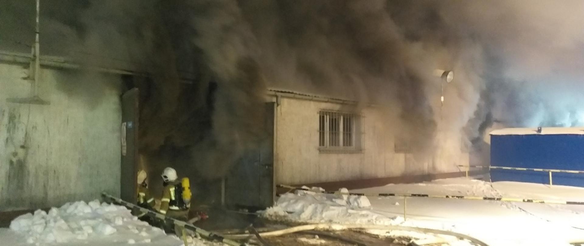 Na zdjęciu przedstawiony jest budynek hali magazynowo-produkcyjnej, z której wydobywa się gęsty, ciemny dym przez otwory okienne oraz otwarte drzwi garażowe. Przed drzwiami na kolanach klęczy dwóch strażaków w ubraniach specjalnych, z założonymi hełmami na głowę oraz zabezpieczonych w linię gaśniczą. Za nimi rozwinięta jest linia zasilająca składająca się z węża pożarniczego. Strażacy na plecach mają założone aparaty ochrony dróg oddechowych. Na zewnątrz, przed budynkiem zalega warstwa śniegu.