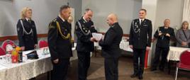 Druhowie oraz komendant miejski państwowej straży pożarnej stoją w świetlicy remizy. Burmistrz wręcza druhowi decyzję