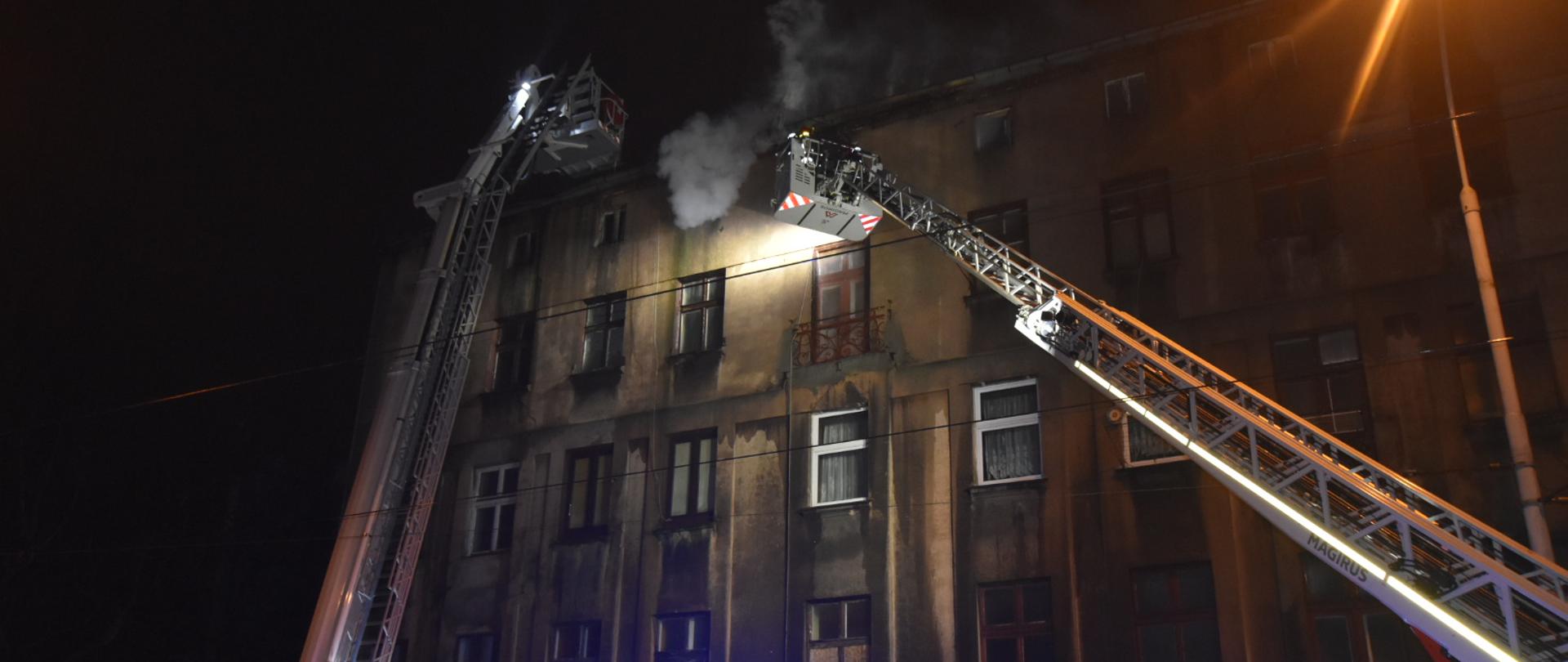 Na zdjęciu dwie drabiny mechaniczne na tle kamienicy, z których strażacy gaszą pożar poddasza.