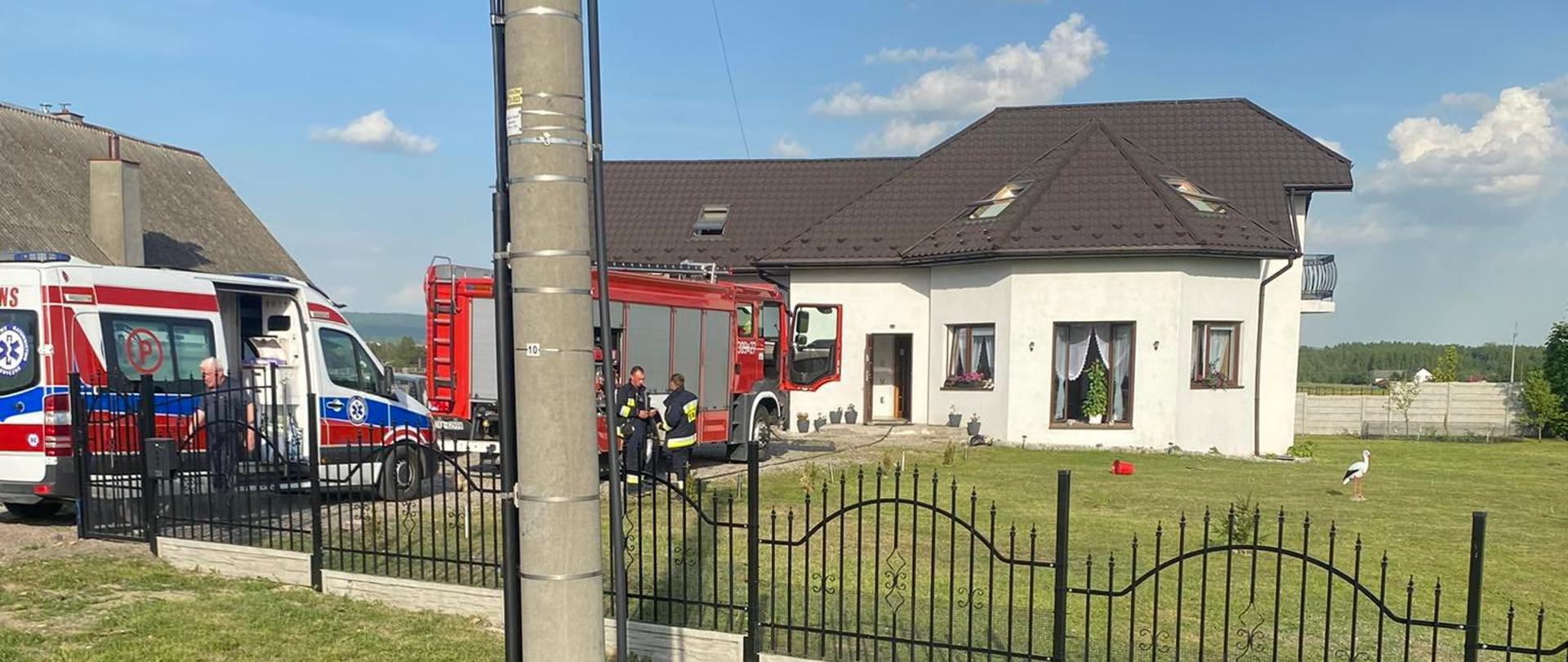 Fotografia przedstawia budynek mieszkalny, w którym wybuchł pożar. Przed budynkiem stoi zaparkowany samochód pożarowy i karetka Zespołu Ratownictwa Medycznego. 
