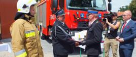 Zdjęcie przedstawia Zastępcę Lubuskiego Komendanta Wojewódzkiego Państwowej Straży Pożarnej, który przekazuje akt wręczenia nowego samochodu naczelnikowi OSP Strużka na tle nowego samochodu oraz strażaka w umundurowaniu specjalnym.