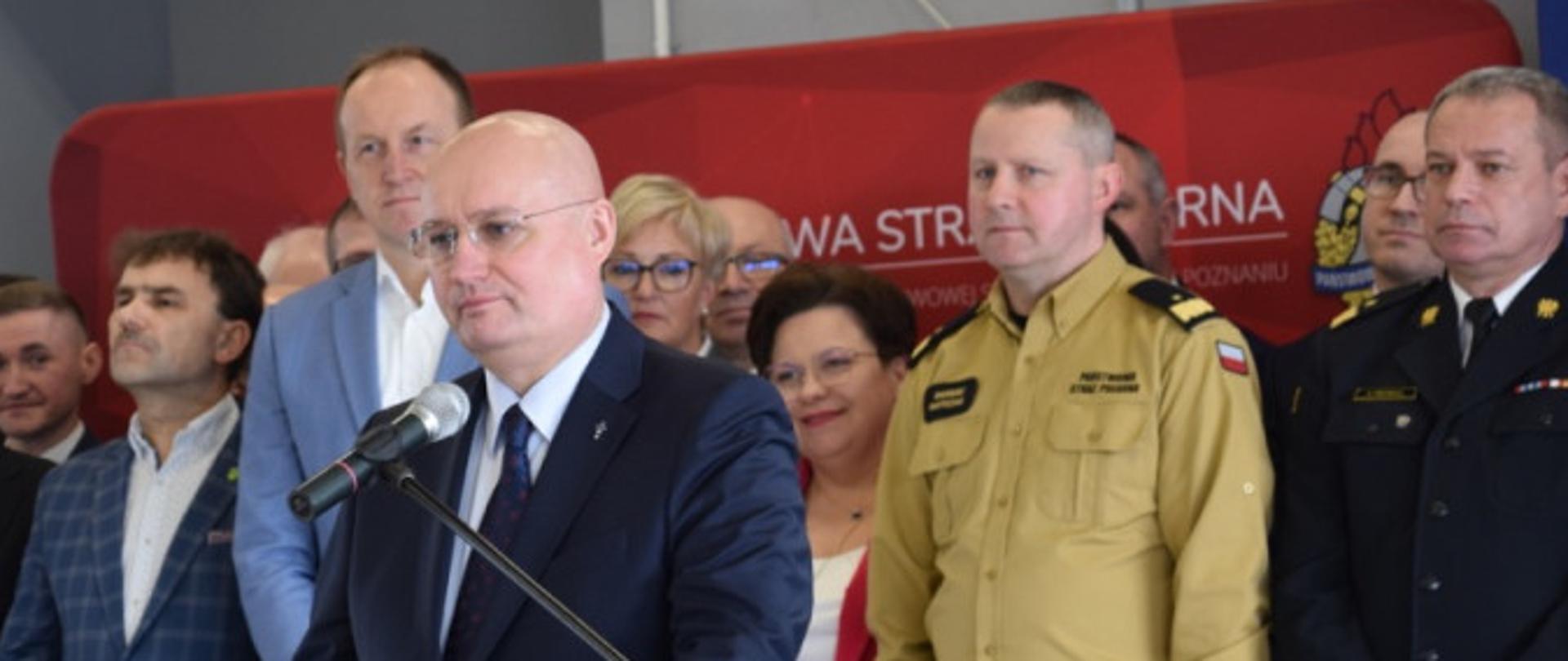 duża grupa kobiet i mężczyzn stoi przed czerwonym banerem, przed nimi mężczyzna w okularach i garniturze stoi przy mównicy