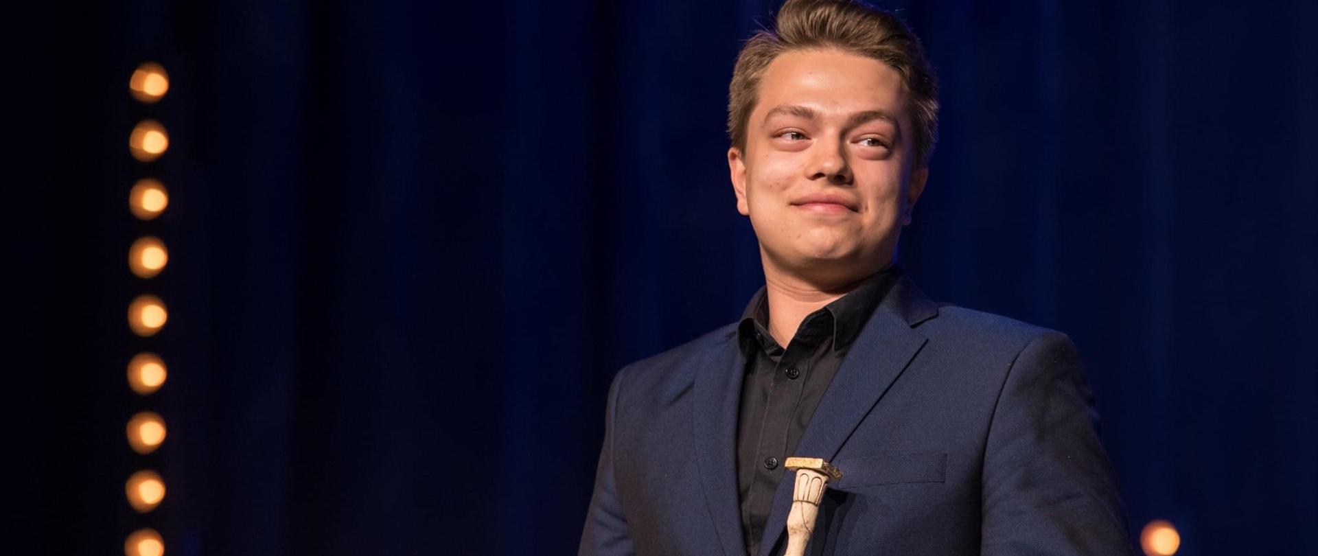 Laureat trzyma w ręku nagrodę, jest ubrany w ciemny garnitur