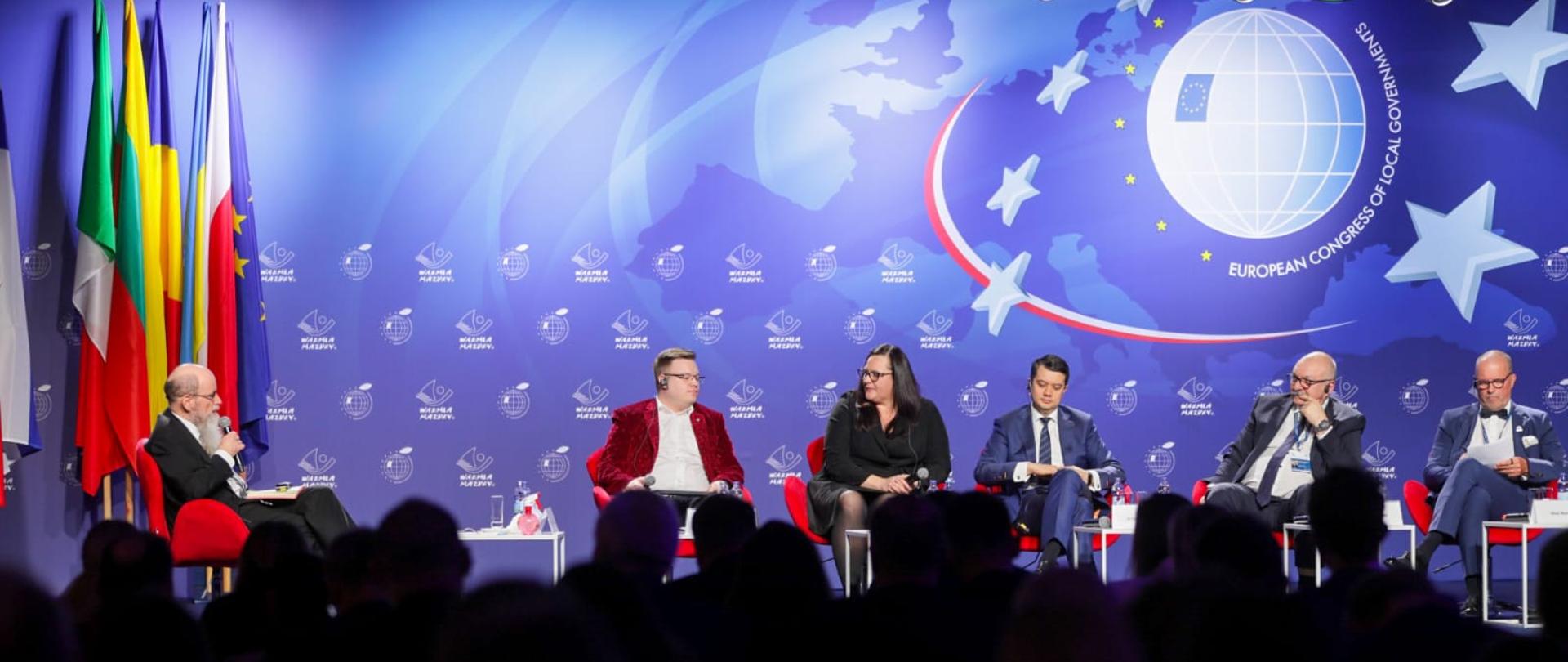 Sześć osób siedzi obok siebie na fotelach na scenie. W środku wiceminister funduszy i polityki regionalnej Małgorzata Jarosińska-Jedynak.