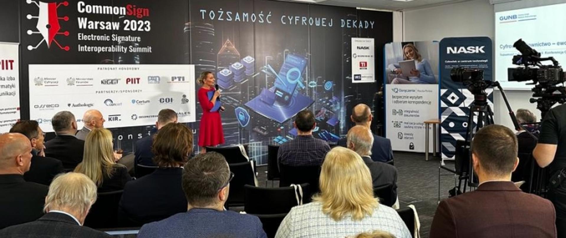 Konferencja CommonSign Warsaw 2023 z prelegentką na scenie, banerem „Tożsamość cyfrowej dekady” i logotypami partnerów; widoczna publiczność i operator kamery.