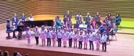 Około 15 dzieci w tych samych kolorowych strojach stoją w rzędzie i śpiewają, za nimi gra orkiestra, po lewej stronie dziewczyna gra na fortepianie.