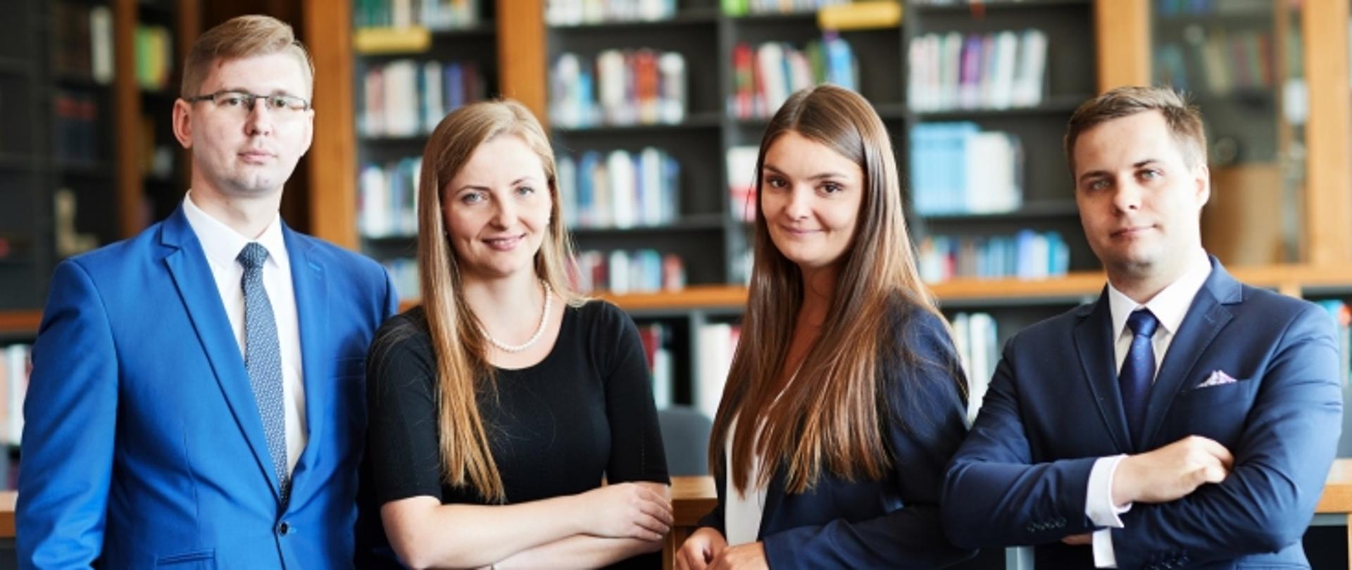 Na tle półek z książkami stoją cztery osoby. Są to dwie elegancko ubrane, uśmiechnięte kobiety i dwaj mężczyźni w garniturach. 