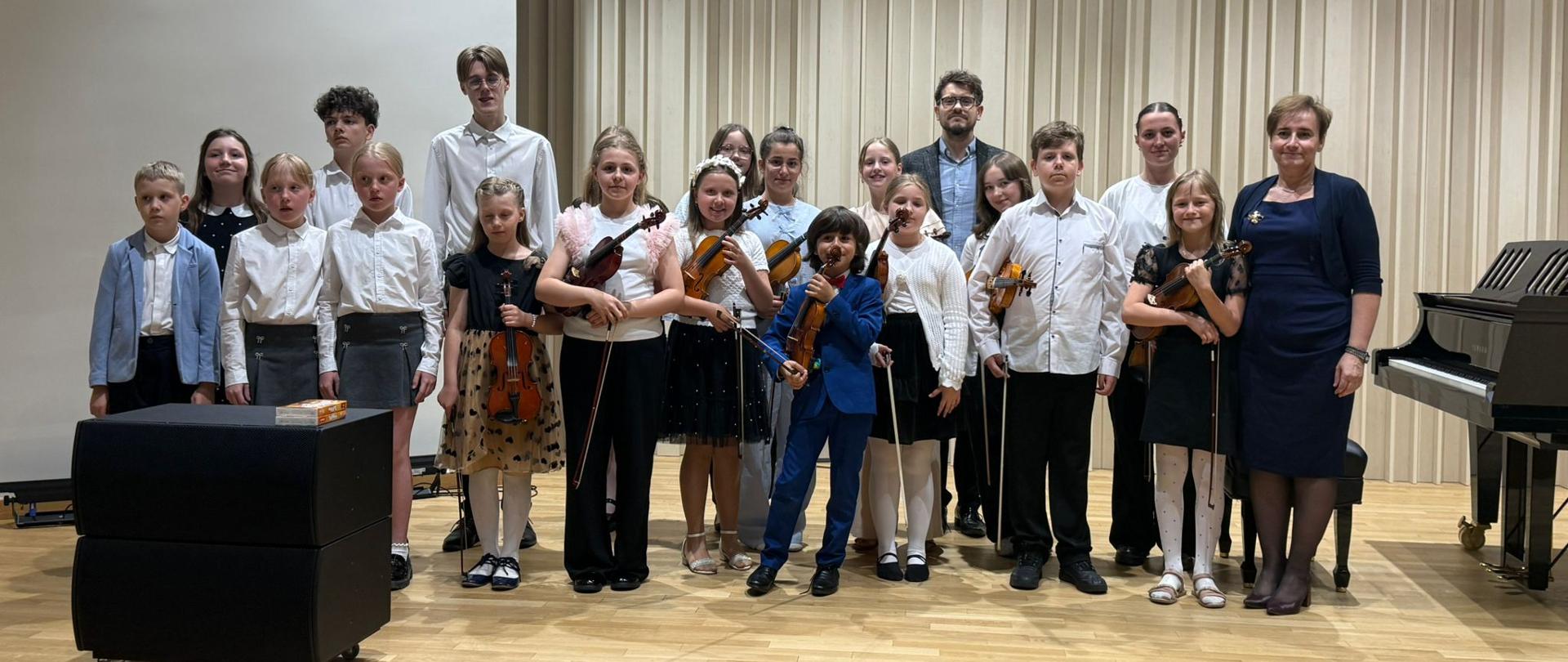 Na zdjęciu duża grupa młodych artystów, którzy wystąpili podczas koncertu uczniów klasy skrzypiec i klasy fortepianu. Wszyscy w odświętnych strojach. Część z nich trzyma skrzypce. Wśród młodych artystów kobieta szatynka w granatowej sukni oraz mężczyzna w niebieskiej koszuli i szarej marynarce. Scena i ściany w odcieniach beżu. 