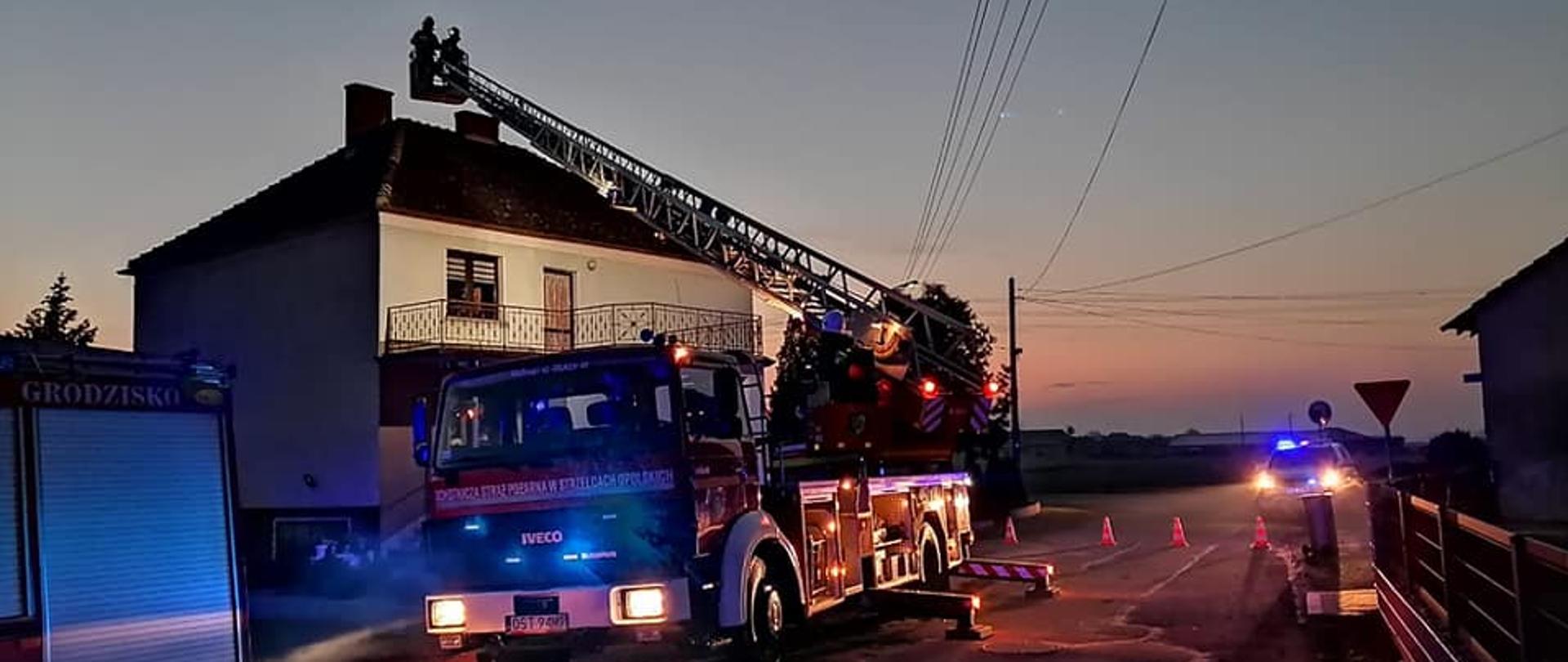 Zdjęcie przedstawia drabinę strażacką z dwoma ratownikami w koszu, którzy przystępują do gaszenia pożaru sadzy w kominie.