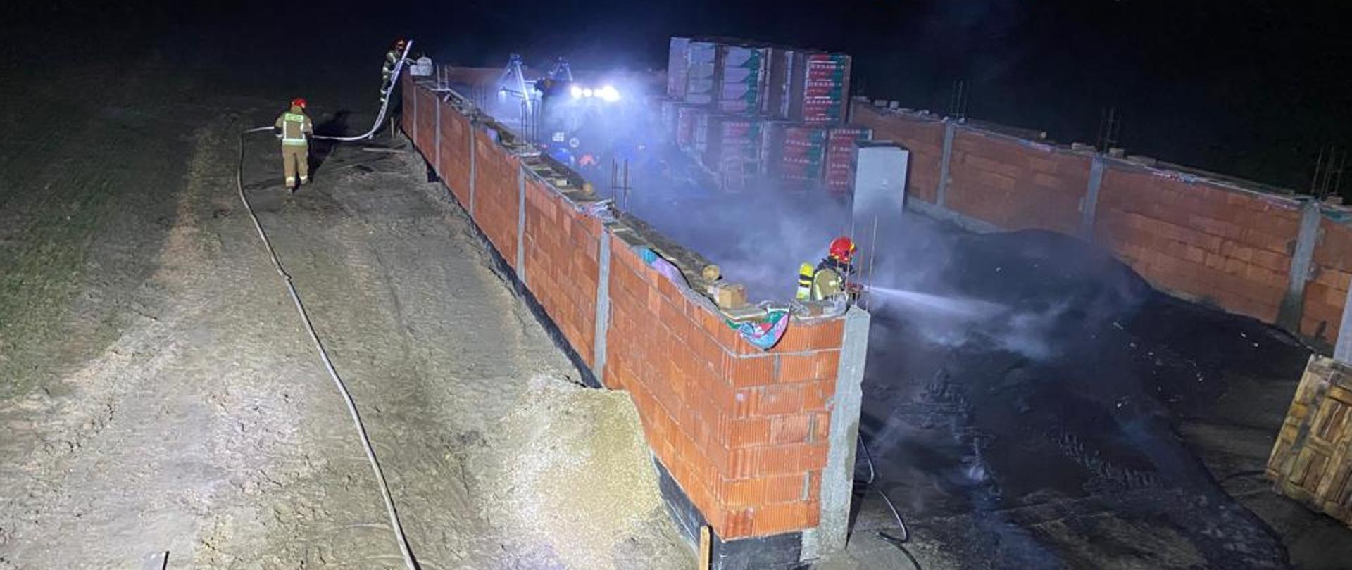Zdjęcie przedstawia rozpoczętą budowę magazynu. W środku znajduje się nawóz organiczny, z którego wydobywa się dym. Jeden ze strażaków w ubraniu specjalnym oraz aparacie dróg oddechowych gasi tlący się materiał. Pozostali dwaj strażacy nadzorują pożar z drugiej strony budynku przy użyciu drabiny przystawnej.