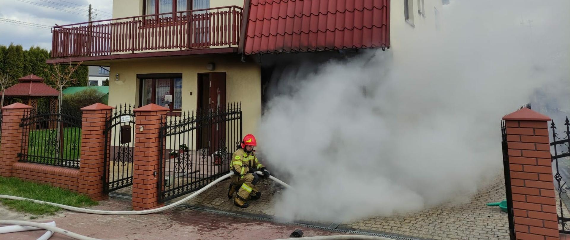 Budynek mieszkalny jednorodzinny, z garażu wydobywa się dym, w linii ogrodzenia kuca strażak ubrany w ubranie specjalne i sprzęt ochrony układu oddechowego, w rękach trzyma linię wężową