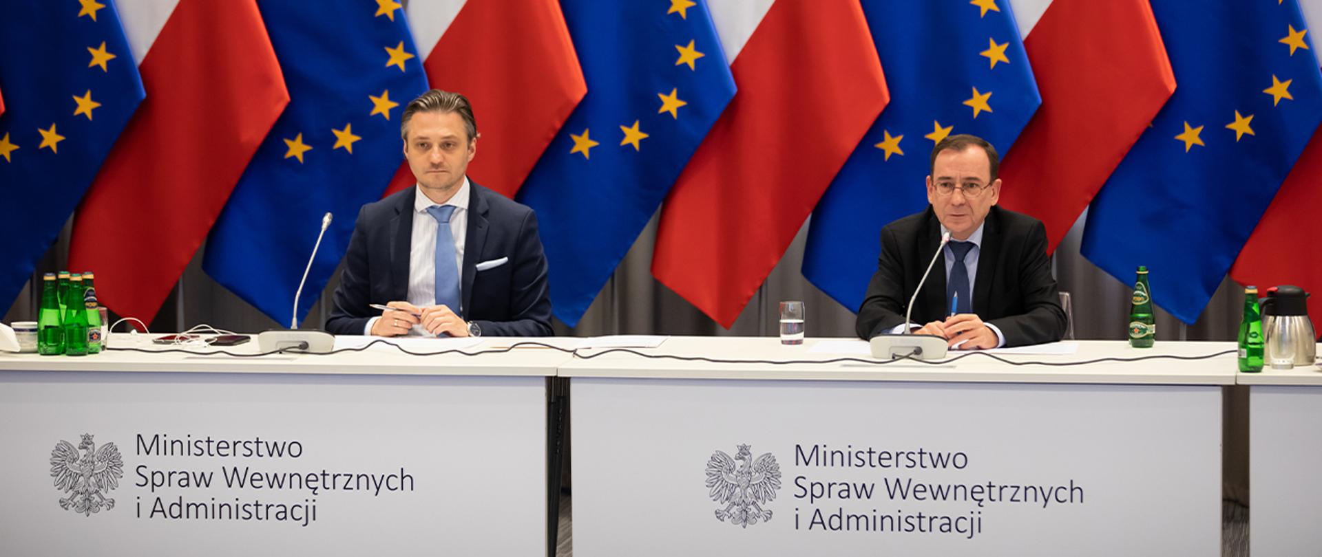 Na zdjęciu widać Mariusza Kamińskiego ministra SWiA i Bartosza Grodeckiego wiceministra SWiA siedzących za stołem w trakcie wideokonferencji. W tle widać rząd flag narodowych i UE ustawionych naprzemiennie.