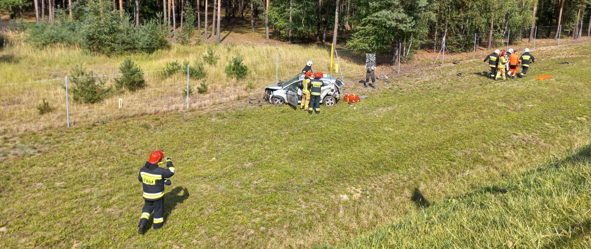 Dzień. Przy ogrodzeniu autostrady stoi rozbity samochód osobowy, strażacy z ratownikami medycznymi transportują na noszach osobą poszkodowaną do ZRM.