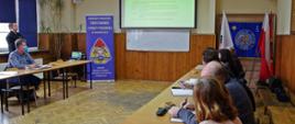 Widok sali szkoleniowej. Przedstawiciele urzędów miast i gmin siedzą przy stolikach i patrzą na ekran widoczny na drugim planie. Obok rolap z logo KP PSP w Wadowicach.