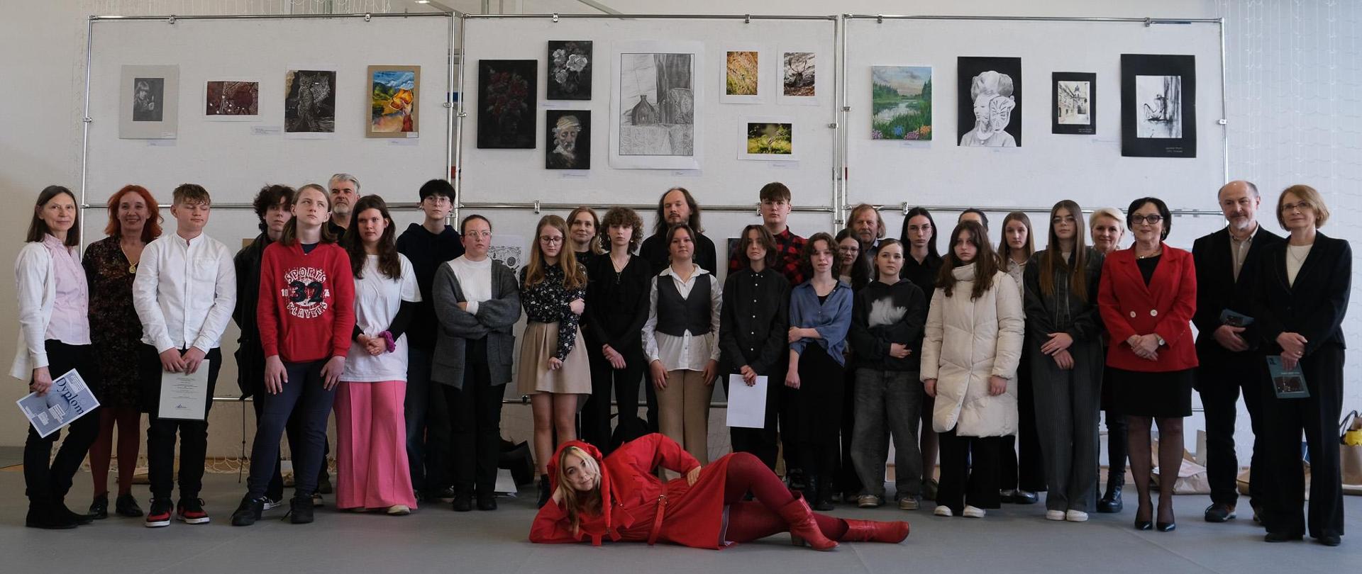Duża grupa osób pozuje do zdjęcia na tle eksponowanych prac. Na pierwszym planie leży podpierając głowę osoba przebrana za Stańczyka