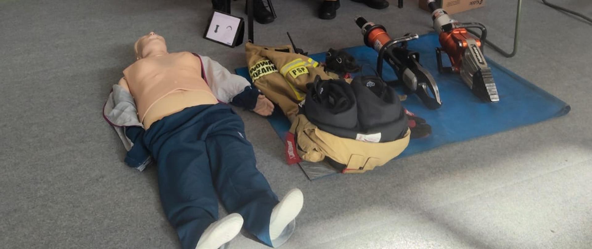 Dwóch strażaków stoi przy stoliku podczas prezentacji sprzętu ratowniczego; na podłodze leżą fantom do nauki resuscytacji, elementy umundurowania i narzędzia hydrauliczne