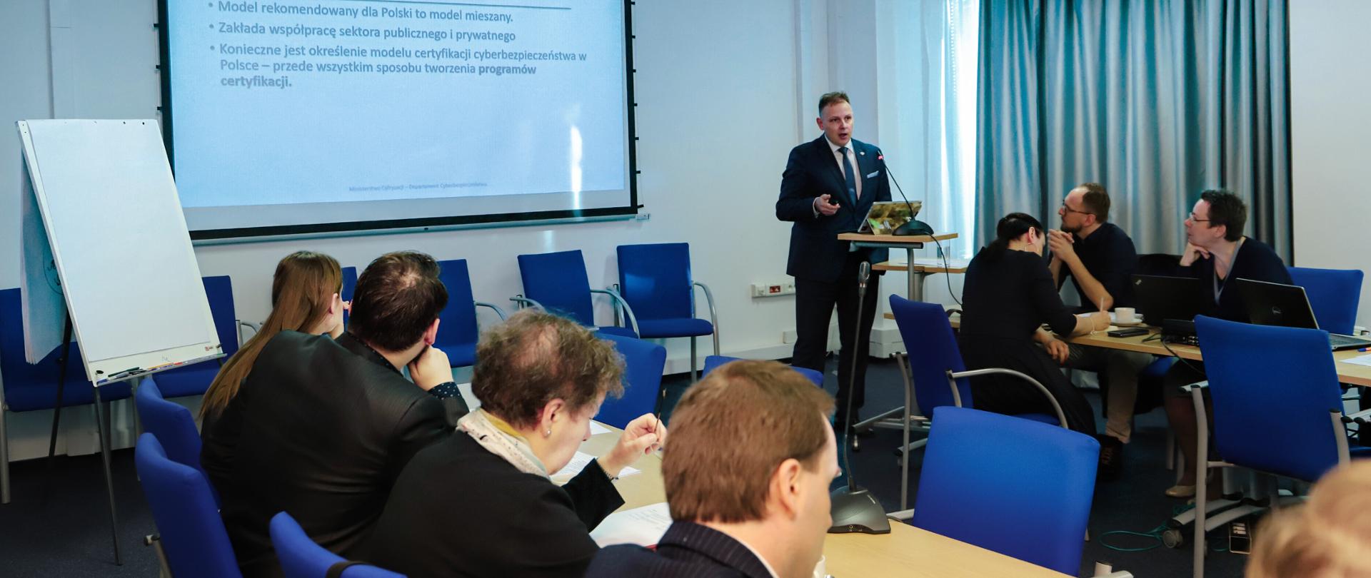 Sala konferencyjna. Dyrektor Robert Kośla przedstawia prezentację, za nim wisi ekran. Przy stolikach siedzą uczestnicy spotkania