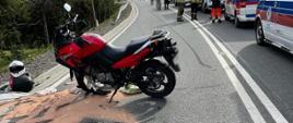 Motocykl na drodze a obok rozlane płyny eksploatacyjne. Na poboczu stoją samochody służb ratowniczych.