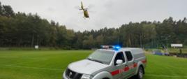 Samochód strażacki lekki stojący na płycie boiska z włączonymi światlami błyskowymi, w tle startujący śmigłowiec pogotowia ratunkowego 